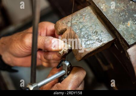 Homme joaillier Goldsmith travaillant dans son atelier de fabrication de bijoux créant et sciant un bijou en or blanc ou en argent dans son atelier de travail. Banque D'Images