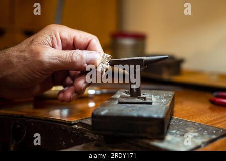 Homme joaillier Goldsmith travaillant dans son atelier de fabrication de bijoux créant et réparant un bijou en or blanc ou en argent avec une enclume dans son atelier de travail. Banque D'Images