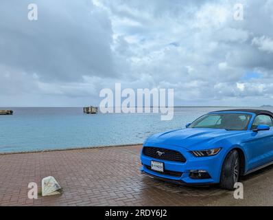 Une voiture Ford Mustang bleu vif se trouve sur le front de mer humide à Kralendijk, Bonaire, Antilles sous le vent avec une vue sur les Caraïbes vertes et bleues. Banque D'Images