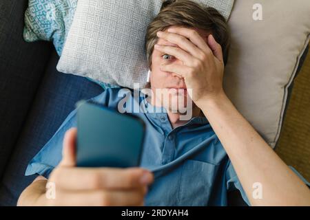 Homme inquiet regardant le téléphone intelligent allongé sur le lit Banque D'Images