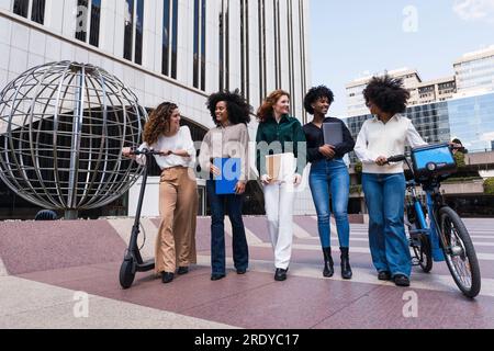 Femmes d'affaires heureuses avec vélo électrique et scooter de poussée marchant dans le parc de bureaux Banque D'Images