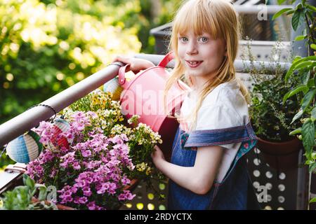 Fille souriante avec arrosoir debout à côté du pot de fleurs dans le balcon Banque D'Images