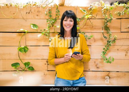 Femme d'affaires heureuse debout avec téléphone intelligent et carte de crédit devant le mur Banque D'Images