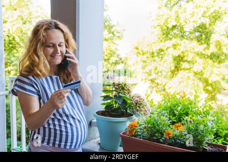 Femme enceinte souriante tenant la carte de crédit et parlant sur le téléphone intelligent Banque D'Images