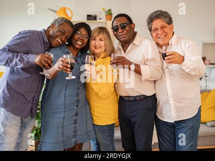 Heureux amis seniors multiculturels tenant des verres à vin debout à la maison Banque D'Images