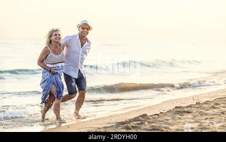 Heureux couple d'âge moyen en amour marchant dans l'eau sur la plage au coucher du soleil. Banque D'Images