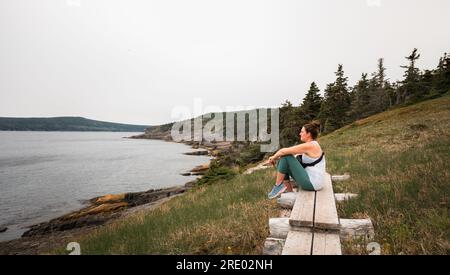 Femme assise sur un chemin en bois regardant l'océan sur une randonnée. Banque D'Images