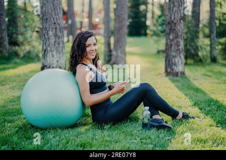 Femme brunette satisfaite assise contre une grosse balle de fitness, en utilisant son téléphone pour la musique, sur l'herbe verte dans une forêt ensoleillée. Concept sportif, porter des sports Banque D'Images