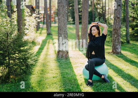 Femme de fitness réfléchie est assise sur le ballon de fitness dans la forêt verte, portant un t-shirt noir et des leggings. Bras levés au-dessus de la tête, contemplant tout en appréciant Banque D'Images