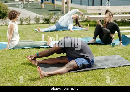 Homme actif afro-américain faisant l'exercice d'étirement tout en étant assis sur le tapis sur la pelouse verte du terrain de sport contre groupe de sportives Banque D'Images