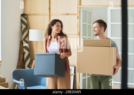 Heureux jeune mari et femme qui se regardent tout en portant des boîtes en carton emballées lors de la réinstallation dans leur nouvel appartement Banque D'Images