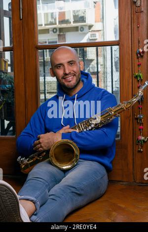 portrait d'un jeune homme latino avec barbe et ongles saxophoniste assis sur le sol à la maison avec son saxophone regardant la caméra. Banque D'Images