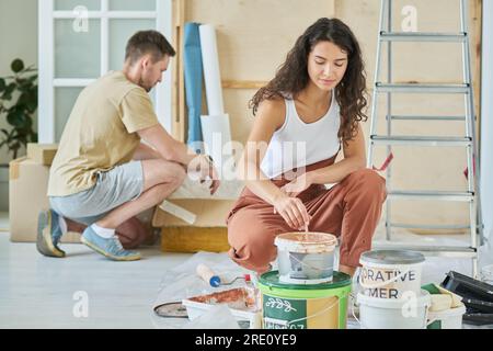 Jeune femme brune en combinaison mélangeant de la peinture liquide beige dans un seau en plastique tout en étant assise sur des squats devant la caméra contre son mari Banque D'Images