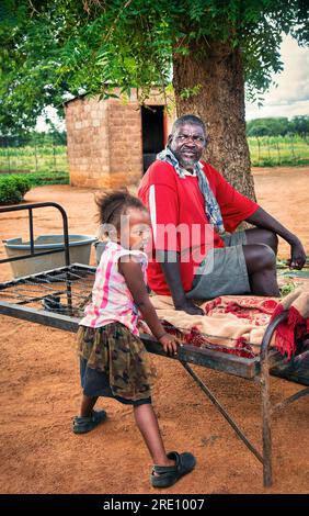 Village africain, vieil homme avec petite-fille situé sur le lit métallique dans la cour ayant un repos à l'ombre d'un arbre Banque D'Images