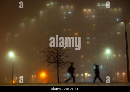 Silhouettes floues de piétons marchant dans la rue avec des lampadaires et un immeuble résidentiel dans un brouillard dense la nuit Banque D'Images
