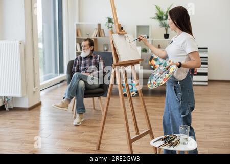 Vue latérale de dame caucasienne dans des vêtements confortables appliquant de la peinture sur toile de coton tout en gardant les yeux sur sitter dans l'espace de studio. Artiste amateur talentueux obtenant de l'expérience en dessinant de l'homme senior calme. Banque D'Images