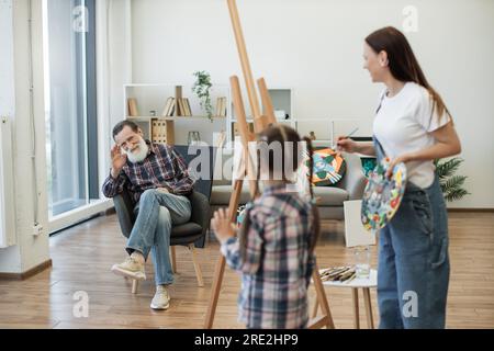 Homme âgé amical en jeans saluant la petite fille debout près de l'artiste féminine avec palette dans le home studio. Souriant retraité dans la chaise douce saluant petite-fille sur la leçon d'art avec la mère. Banque D'Images