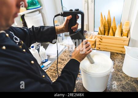 Gros plan moyen de la production de crème glacée. Homme utilisant un blender professionnel pour mélanger la crème glacée. Équipement professionnel et cornets de crème glacée en arrière-plan. Photo de haute qualité Banque D'Images