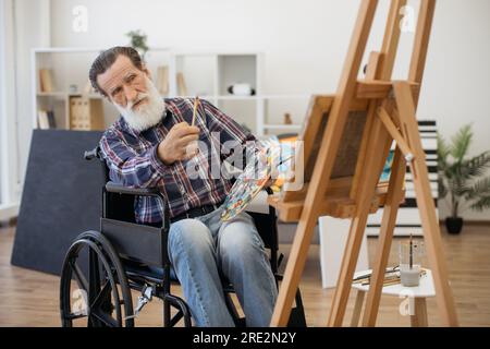 Homme retraité à barbe grise handicapé assis devant un chevalet en bois et examinant un dessin. Homme âgé passant son passe-temps seul à peindre sur toile Banque D'Images