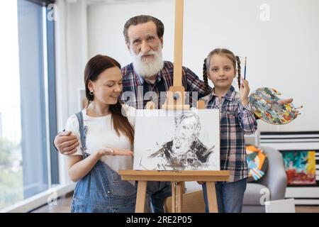 Famille d'artistes embrassant ensemble près de chevalet avec photo à la galerie d'art lumineuse. Grand-père avec petite-fille en chemises à carreaux regardant droit i Banque D'Images