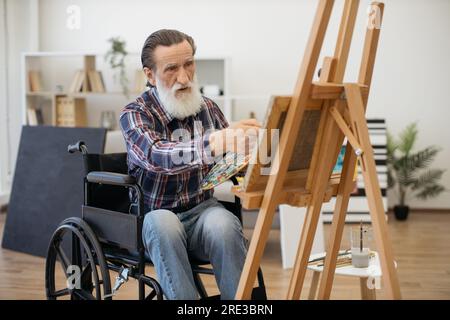 Homme retraité à barbe grise handicapé assis devant un chevalet en bois et examinant un dessin. Homme âgé passant le passe-temps seul à peindre sur toile dans un salon spacieux lumineux. Banque D'Images