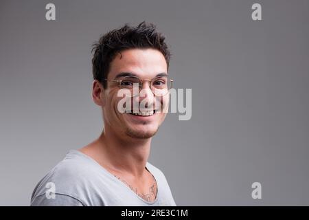 Portrait de trois quarts d'un jeune homme bespectacé. Sa musculation et son regard de tatouage sur sa chemise grise sont évidents. Il sourit, heureux, comme si Banque D'Images