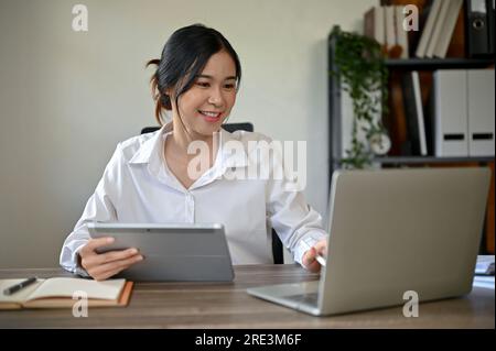 Une femme d'affaires asiatique millénaire professionnelle et attrayante qui gère ses tâches sur sa tablette et son ordinateur portable à son bureau dans le bureau. Banque D'Images