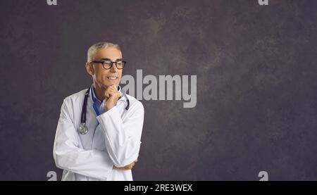 Médecin principal pensif souriant qui rêve ou pense tout en se tenant debout sur fond gris. Banque D'Images