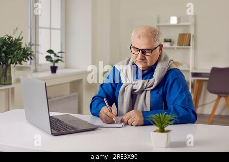 Homme senior travaillant avec un ordinateur portable et prenant des notes sur papier sur le lieu de travail Banque D'Images