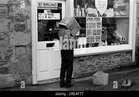 Village shop 1970s Royaume-Uni. Un jeune garçon avec une boîte en carton vide de chips salées prêtes au-dessus de sa tête à l'extérieur d'un magasin de village, épicerie, qui vend 'crème à vendre et par la poste.' 'St Bruno le tabac qui ne sera pas pressé.' Garde les cigarettes. Les offres spéciales des magasins incluent, ' Heinz Salad Cream, carénages de cornouailles, Ambrosia Milk puddings, Gales Lemon Curd, R / Mixed Mustard, Spillers Bonio et Kenomeat et Smedleys Beetroot." Les poupées en plastique pour enfants viennent dans un sac en plastique transparent et accrochent à la fenêtre. Ils vendent également une variété de crèmes glacées. St Just, Cornwall. 1970 Angleterre. HOMER SYKES. Banque D'Images