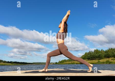 Femme pratiquant le yoga faisant l'exercice Virabhadrasana, pose de guerrier numéro un, s'exerçant en vêtements de sport sur la rive d'un lac par une chaude matinée ensoleillée Banque D'Images