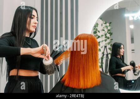 Femme coiffeuse fait peigner les cheveux rouges au client et fait la coiffure dans le salon de beauté devant le miroir. Coiffage et lissage des cheveux. Banque D'Images