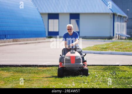 Le jardinier homme à main âgé tond la pelouse sur le tracteur de tondeuse à gazon. Un homme âgé travaille comme ouvrier. Territoire de tonte de l'installation industrielle le jour de l'été. Véritable workflow. Banque D'Images