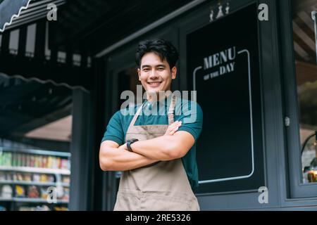 Jeune homme d'affaires asiatique adulte croisant ses bras devant le café restaurant tout en souriant. Heureux et joyeux barista de Coffee Shop ayant les bras croisés. Photo de haute qualité Banque D'Images