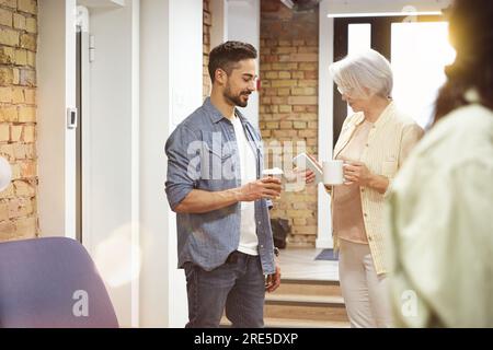 Deux collègues tenant le café, regardant les smartphones tout en se tenant debout dans un espace de bureau moderne. Entreprise, coworking, concept de gestion Banque D'Images