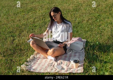 Une jeune femme est assise sur un tapis dans le parc et travaille. Femme avec ordinateur portable dans le parc Banque D'Images