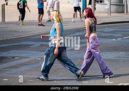 Paire de jeunes femmes minces, filles ou adolescentes, habillées en jeans, marchant à travers une route et faisant du shopping en été, Royaume-Uni. Amis dans les magasins. Banque D'Images