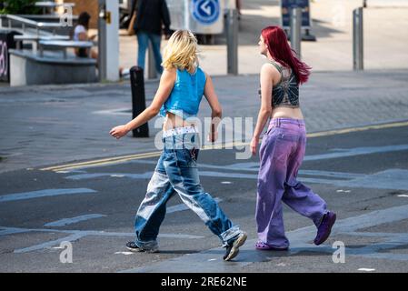 Paire de jeunes femmes minces, filles ou adolescentes, habillées en jeans, marchant à travers une route et faisant du shopping en été, Royaume-Uni. Amis dans les magasins. Banque D'Images