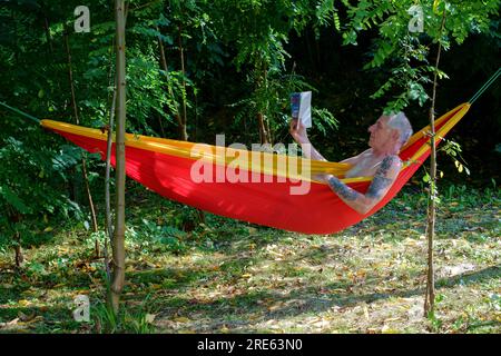 homme plus âgé se relaxant dans le jardin hamac ombragé par les arbres lisant le livre zala comté hongrie Banque D'Images