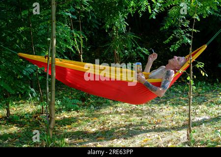 homme plus âgé se relaxant dans le jardin hamac ombragé par les arbres avec cigarette et canette de bière zala comté hongrie Banque D'Images