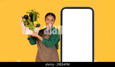 Femme noire près de l'énorme téléphone tenant la boîte de légumes, fond jaune Banque D'Images
