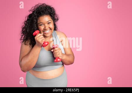 Excité femme noire taille plus en vêtements de sport tenant des haltères et riant, debout sur fond rose, espace de copie Banque D'Images