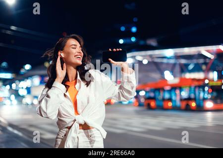 Souriante femme caucasienne utilisant un téléphone portable et des écouteurs tout en marchant dans la ville la nuit, espace libre Banque D'Images