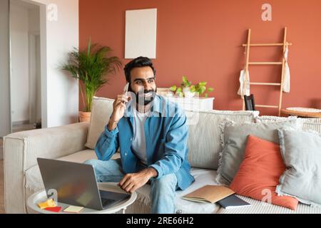 Homme indien ayant une conversation téléphonique agréable assis près de l'ordinateur portable à l'intérieur Banque D'Images