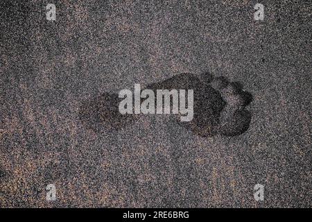 Empreinte unique profonde du pied humain gauche dans le sable volcanique sombre. Banque D'Images