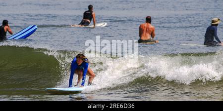 Gilgo Beach, New York, États-Unis - 22 juillet 2023 : les femmes surfent dans un rash Guard bleu à manches longues chevauchant une vague tandis que d'autres s'assoient sur leurs planches de surf en attendant Banque D'Images