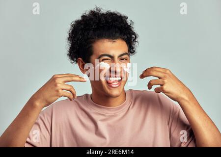 Portrait de studio de beau mec souriant avec les cheveux bouclés noirs appliquant de la crème sur son visage. Joyeux homme swarthy biracial utilise des produits de beauté dans son skinc Banque D'Images