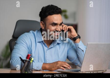 Homme d'affaires indien parlant sur le téléphone mobile tout en travaillant sur ordinateur portable au bureau - concept de conseiller financier, multi-tâches et style de vie d'entreprise Banque D'Images