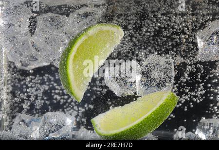 Tranches de citron vert juteux et glaçons dans de l'eau gazeuse sur fond noir, gros plan Banque D'Images