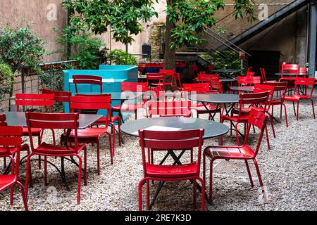 Chaises en métal rouge vif autour de tables grises sur un patio en pierre avec arbres et escaliers. Banque D'Images